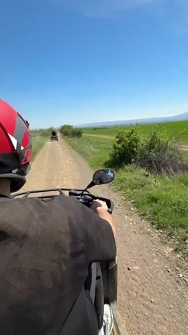 The video shows a first-person view of someone riding a black ATV on a dirt road. The rider is wearing a black shirt, grey pants, and white sneakers.  A red helmet with grey and white accents is on their head. In front of them, two other ATVs are driving along the same road. The environment consists of green fields, sparse bushes, and a distant mountain range under a bright blue sky with some scattered clouds. The road is lined with grass on both sides, and another dirt path runs parallel to the road on the right.