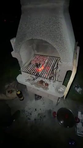 The video shows a nighttime cookout. A concrete barbecue is the main focus, with a grill grate holding several hamburger patties over glowing coals. Off to the side, there is a bag of charcoal, a bottle of beer, and a pot with a lid. A young man in a gray hoodie walks into frame; he appears to be watching the barbecue. There are lights in the distance, possibly from another house.