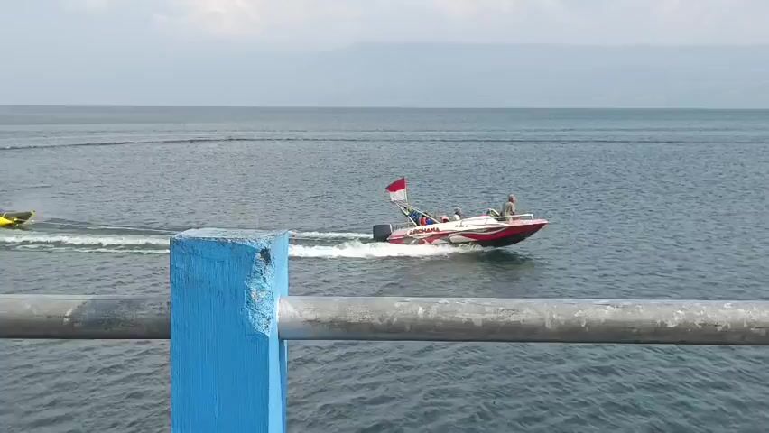 A bright yellow banana boat carrying six individuals in red vests is being towed across a large body of water by a white and red speed boat. The speed boat, with an Indonesian flag attached to its rear, is creating a wake as it moves forward. The water is a deep blue, and the sky is hazy with a distant view of land. In the foreground, a metallic railing with blue vertical supports is visible, suggesting the video is shot from a pier or bridge.