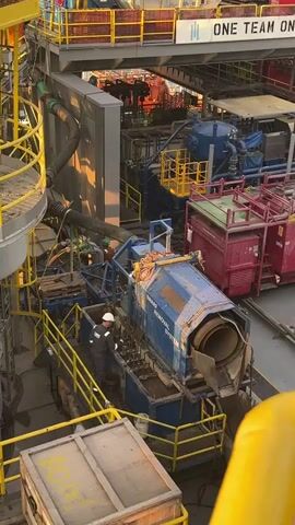 The video shows an industrial setting, possibly an oil rig or a similar offshore platform. In the foreground, a person wearing a hard hat and work clothes is operating a piece of machinery. The machine appears to be a pipe removal system, with pipes being processed and ejected. There are yellow safety railings and structures around the work area, typical of industrial environments. In the background, there are other pieces of heavy machinery, cranes, and buildings, suggesting a complex operational facility. The overall scene is one of industrial activity, with a focus on the operation of specialized equipment and the presence of workers.