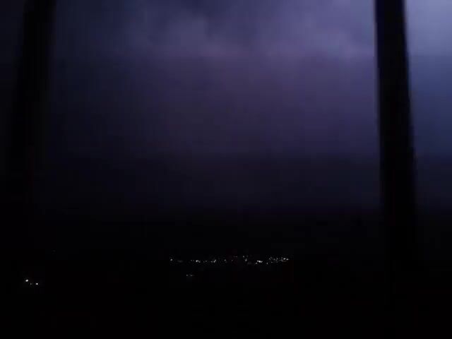 The video depicts a nighttime scene with a distant cityscape. The scene is extremely dark, with only faint lights visible from the city in the far distance. A brief flash of lightning illuminates the sky.