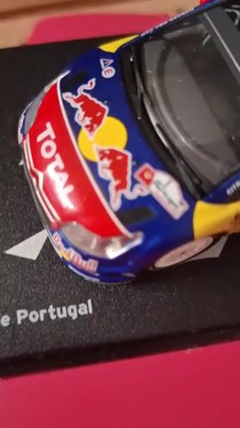 A close-up, overhead shot shows a collectible toy car with a red, white, and blue livery, adorned with logos from Red Bull and Total. The car is positioned on a black base with some text on it, including "Portugal" and "Citroën C4" with "Sébastien Loeb - Dani Sordo". The background is a blurry pink surface. The toy car appears to be a rally car, possibly a Citroën C4 WRC driven by Sébastien Loeb.