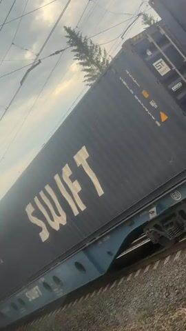 A close-up video shows a train carrying shipping containers. The containers are dark blue with white lettering that reads "SWIFT". The train is moving on a track, and the camera is positioned at an angle that makes the scene appear tilted. The sky is cloudy and the scene is overcast. There are power lines and poles visible above the train. The video is 4 seconds long.