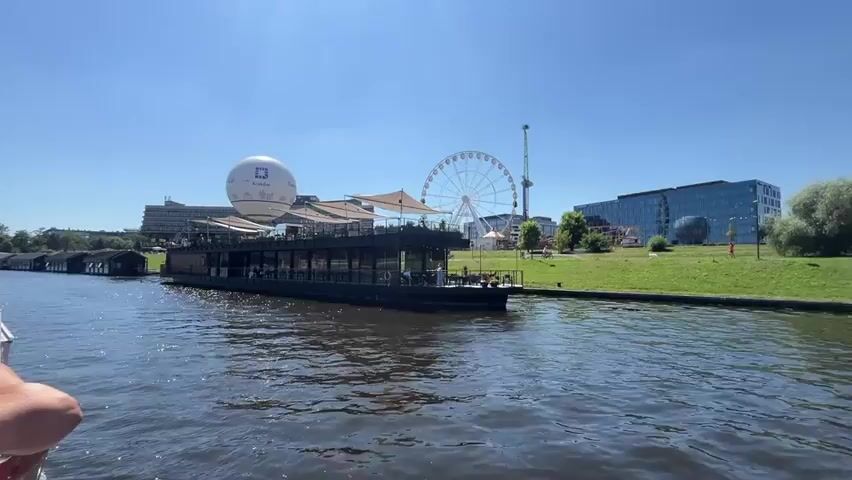 The video captures a view from a boat on a body of water, moving towards a large, dark-colored barge or restaurant with an outdoor seating area. Behind the barge, a large white balloon with a logo is visible, as well as a Ferris wheel and several modern buildings. The sky is clear and blue, indicating a sunny day. Several people are visible on the barge, seated at tables. On the boat from which the video is filmed, the backs of several people's heads and shoulders are visible in the foreground. The water is calm with some ripples.