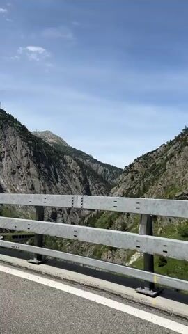 This is a static shot from a vehicle moving along a road. In the foreground, a silver metal guardrail runs horizontally across the frame. Beyond the guardrail, a steep, rocky mountain face dominates the left side of the image, with patches of green vegetation and trees clinging to its slopes. A narrow road or path can be seen winding its way along the base of the mountain on the left side. To the right of the mountain, the landscape opens up into a valley, with another, less steep, tree-covered mountain visible in the distance. The sky above is a clear blue with a few scattered white clouds. The lighting suggests it is daytime, likely late morning or early afternoon.