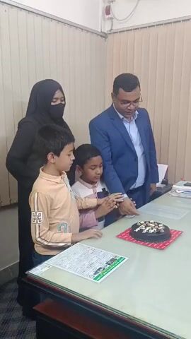Here's a concise description of the video:

Inside what appears to be an office, a man in a blue suit stands with a woman in a black hijab and two young boys around a desk. On the desk is a chocolate cake on a red square. The cake is decorated with white frosting. The man is indicating something on the cake, possibly showing the boys where to cut. There's a document on the desk as well. The room seems to be lit by overhead fluorescent lights.