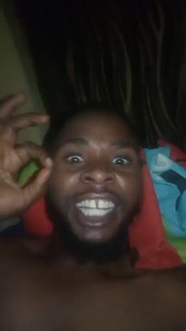 A man with dark skin and a beard is shown from the chest up, looking directly into the camera. He is making an "okay" sign with his fingers. He appears to be indoors, perhaps lying in bed, with a patterned curtain behind him. A red and blue cloth is also visible. He is speaking.