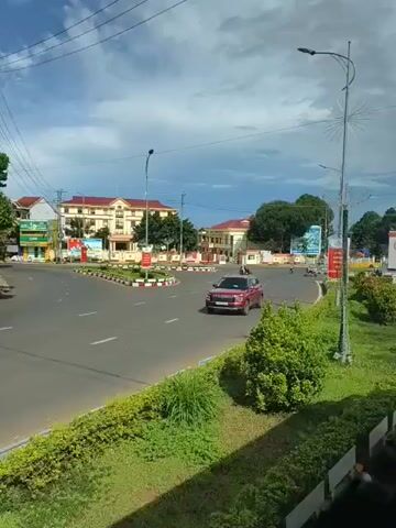 The video pans across a circular intersection in a town. A red SUV is driving through the intersection. There are several buildings in the background, as well as streetlights and decorative poles with flags and banners. The intersection is surrounded by green grass and bushes. The sky is cloudy. There are a few people on motorbikes visible in the distance.