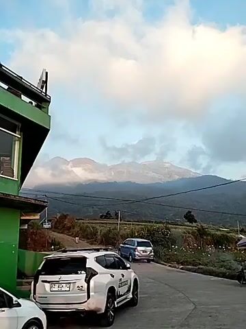 A white SUV is parked on the left side of a paved road. A blue minivan is driving away from the camera down the road. In the background, a mountain range is visible, with clouds obscuring the peaks. The sky is partly cloudy and the lighting suggests it is either early morning or late afternoon. There is some greenery and vegetation on the side of the road.