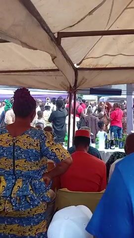 The video shows a crowd of people gathered under a tent, likely at an event. The camera is positioned behind a row of seated individuals, offering a view of the activity taking place further ahead. 

A man in a black shirt can be seen holding a phone, possibly recording or taking pictures of the scene. Several other people are milling around, some dressed in traditional attire. There are also men in red shirts who appear to be acting as security.

A table in front of the seated individuals is laden with drinks, including beer and champagne. The setting suggests a festive or celebratory occasion, possibly a wedding or a similar social gathering.