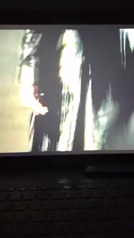 The video shows a laptop screen displaying footage of a music video. The opening scene shows multiple cars on a city street at night, followed by a shot of people walking in leather attire. The final shot depicts a woman with curly red hair yawning. The video is filmed at night and the room is dark, with only the light from the laptop illuminating the scene. The focus is on the laptop screen and the video playing on it.