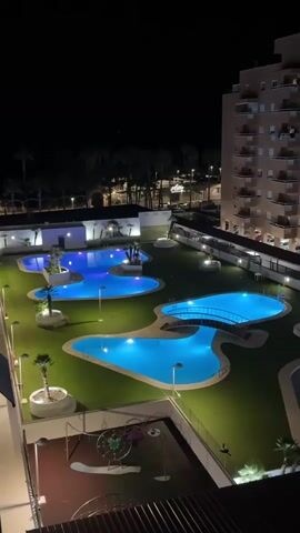 This aerial, high-angle, vertical video captures a nighttime view of an apartment complex's amenities in Les Amplaries, Spain. The scene is dominated by several irregularly shaped swimming pools, illuminated by bright blue lights, creating a vibrant and inviting atmosphere. The pools are surrounded by lush green artificial turf, with some palm trees and other vegetation adding to the tropical feel.

In the foreground, a playground area is visible, featuring a slide and other equipment, also illuminated by artificial light. In the background, a tall, multi-story apartment building stands prominently, its windows lit from within, suggesting a residential setting. Beyond the complex, palm trees line a street where car headlights and streetlights are visible, indicating it's a populated urban or resort area. A sign with "Atlas" is partially visible above the street.

The time of day is clearly night, with the sky being a deep black, and the artificial lighting playing a crucial role in illuminating the scene. There is no indication of weather conditions other than the darkness. The overall mood evoked is one of tranquility and leisure, with the glowing pools and well-maintained grounds suggesting a vacation or resort destination. No people are visible in the video.