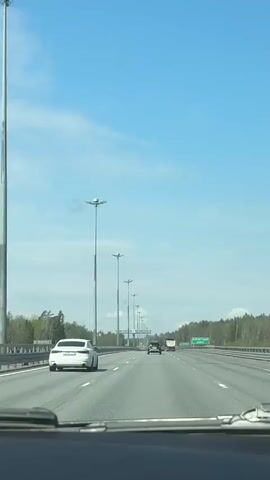 The video captures a car's perspective while driving down a highway. The sky is blue with scattered clouds. On the road, there are several other vehicles: a white sedan in the closest lane, a black SUV further ahead, and a box truck in the distance. Tall lampposts line the road with a unique multi-bulb design. A green highway sign indicates "КУРОРТНЫЙ РАЙОН" and "р. ЧЕРНАЯ". The surrounding environment includes trees bordering the highway on both sides.