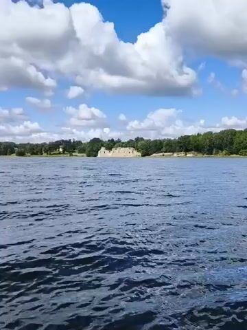 The video shows a wide shot of a body of water with a ruin in the distance. The water is dark blue and has a rippling texture, suggesting it is being disturbed by wind or movement. The sky above is bright blue with scattered white, puffy clouds. The ruin appears to be an old stone structure, possibly a castle or abbey, with several walls still standing but lacking a roof. It is situated on a gently sloping bank covered in green trees and vegetation. The overall scene depicts an outdoor environment on a clear day, likely during daylight hours. There are no people or discernible objects in the foreground or mid-ground. The camera appears to be mounted on a moving platform, as the perspective of the ruin shifts slightly throughout the short video clip.