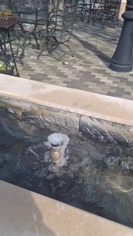 The video shows a tranquil outdoor scene. A narrow water fountain with a small bubbling fountainhead is featured in the foreground, its basin lined with dark, marbled stone. The fountain's edges are constructed from light-colored, possibly limestone, slabs. The camera pans to the right to reveal a decorative stone planter filled with pansies, its colors including yellow, purple, and black. The planter sits on the edge of the fountain structure. In the background, there is a patio area with ornate wrought-iron chairs and a paved ground. A black, cylindrical bollard is also visible, along with partially obscured furniture. The lighting suggests it is a sunny day.