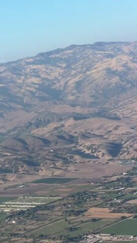 The video shows an aerial view of a landscape with rolling hills and agricultural fields. In the foreground, there are patches of green fields and some buildings. Further back, the landscape transitions to dry, brown hills with some sparse vegetation. The sky is clear and blue. The camera is in motion, giving the impression of flight. There are no people or specific actions clearly visible beyond the movement of the camera.