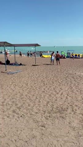 The video captures a crowded beach scene on a sunny day. The camera pans across a sandy beach, showing numerous people relaxing under umbrellas, swimming in the turquoise water, and engaging in various beach activities. In the foreground, two men are seen standing on the sand, facing each other, as if in conversation or about to interact. Further back, several inflatable water toys and a boat are visible in the water. The overall atmosphere appears to be one of leisure and recreation, with people enjoying the summer weather and the beach environment. The sky is clear and blue, indicating good weather conditions.