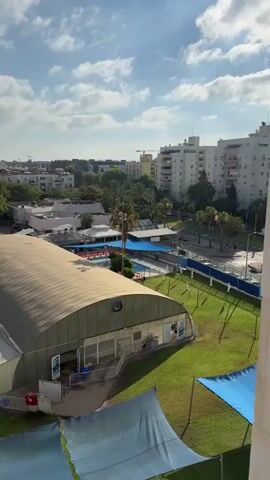 The video displays an aerial view of a complex featuring a large, arched building with a tan roof and white walls, appearing to be an indoor sports facility or gymnasium. In front of the building, a well-maintained green lawn is visible, with several large, blue shade structures spread out across it. Beyond the lawn and the arched building, a swimming pool area with lounge chairs can be seen. The background shows a cityscape with various buildings, trees, and a cloudy blue sky. The camera pans slightly to the left, offering a broader perspective of the surroundings.