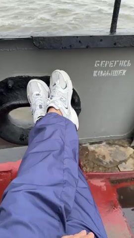 The video opens with a close-up shot of a person's feet wearing white sneakers and blue pants. The person is sitting on a boat, with their legs extended. The background shows a grey, choppy body of water and the railing of the boat. The camera then pans out to reveal more of the boat's railing and the vast expanse of water. A distant shoreline with trees is visible. The sky is overcast and grey, suggesting a cloudy day. The overall atmosphere is calm and serene, with a focus on the journey across the water. There is a warning sign on the boat that reads "БЕРЕГИСЬ КОЛЬШЕК" (Beware of bumps).