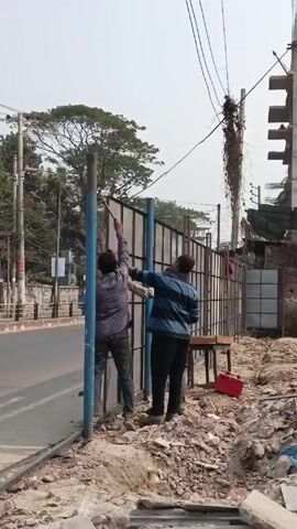 The video shows scenes from a construction site next to a busy road. Two men are installing a metal fence. There is rubble and debris on the ground. Another man and woman are working on a pile of bricks. A partially-constructed building is visible in the background. The video pans to show a long, white wall, possibly a fence. Finally, it shows a sidewalk with pedestrians walking by the construction site. There are shops and street signs visible in the background.