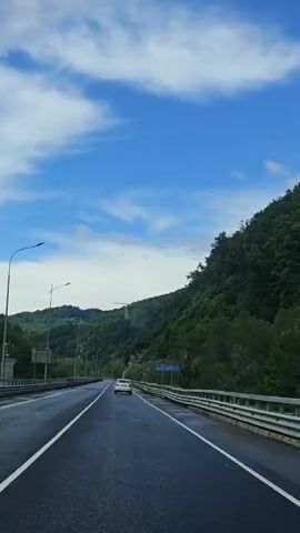 The video shows a view from a car driving on a highway in a mountainous area. The sky is blue with scattered white clouds. On either side of the highway are tree-covered mountains. A sign indicates "р.Мзымта". A cell tower is visible atop one of the mountains.