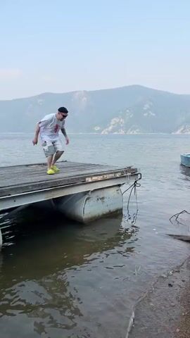A man wearing a black beanie, sunglasses, a white t-shirt with an orange graphic, and yellow Crocs runs from a dock into the water. He lands on his feet and then squats down at the water's edge. He then stands up, smiles at the camera, and walks away from the water onto a rocky shore. In the background, there is a lake with mountains and boats.