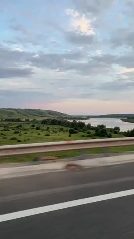 This is a low-angle, medium shot of a landscape viewed from the side of a moving vehicle. The foreground shows a highway with a white line dividing the lanes, and a guardrail separates the highway from the natural scenery. The natural scenery includes rolling green hills dotted with trees, and a body of water, likely a river or lake, in the distance. The sky is partly cloudy with a soft, diffused light, suggesting either early morning or late evening. The overall impression is one of travel and a picturesque natural environment.