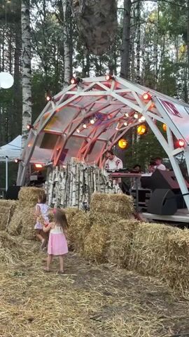 The video shows an outdoor event with a stage set up in a wooded area. Two young girls, one in a pink dress and the other in a white and pink dress, are walking towards the stage. The stage itself is covered by a white arched structure with colorful lights. There is a DJ booth with at least two people working. In front of the stage, there are stacks of hay bales, and the ground is covered in dry grass and leaves. In the background, there are trees, and a large, abstract, mask-like decoration is hanging from one of the trees above the stage. The overall atmosphere appears festive and casual, possibly a music festival or outdoor party.
