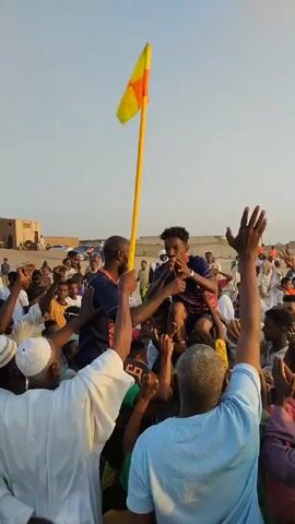 Here is a concise description of the video:

A crowd of people is gathered in an open area, likely a field or rural setting, under a bright, clear sky. At the center of the crowd, two men are being hoisted up, one of whom is holding a long yellow pole with a yellow and orange triangular flag on top. The crowd appears to be celebrating or cheering, with many individuals raising their arms in the air. Some people are wearing traditional white clothing, while others are in more casual attire. In the background, there are some buildings and parked cars visible, suggesting a nearby village or town.