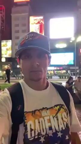 The video features a man recording himself in a brightly lit urban environment at night. He is wearing a white t-shirt with a colorful graphic, a blue and red baseball cap, and a black backpack. The camera is positioned at chest height, indicating that the man is holding it. He appears to be walking or standing in a plaza with many people around. In the background, there are several tall buildings illuminated with bright lights and digital billboards displaying advertisements. The scene suggests a bustling city center with a vibrant nightlife.