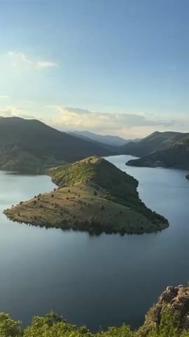 The video shows a wide, calm lake surrounded by rolling, tree-covered hills under a clear blue sky with scattered clouds. A small boat is visible on the lake, leaving a wake behind it. The foreground is a rocky, overgrown area, suggesting a viewpoint from a cliff or high ground. The sun appears to be low in the sky, casting a warm light on the landscape.