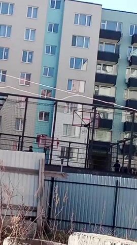 The video shows an outdoor basketball court in an urban setting. There are at least three people on the court, appearing as small figures from the camera's perspective. The court is enclosed by a black wire fence. Beyond the fence is a gray corrugated metal fence and some scattered debris and dry vegetation. In the background, there is a tall multi-story apartment building, with its facade featuring a combination of gray, light blue, and white sections. The building has numerous windows, some with visible curtains or personal items inside. The sky is clear and light in color. The camera is relatively static, providing a view through the fence towards the basketball court and the building in the background.