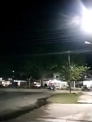 The video is a night-time recording of a street. A bright streetlight illuminates the scene. In the background, there are buildings and trees. A vehicle is seen driving on the street. The camera is stationary.