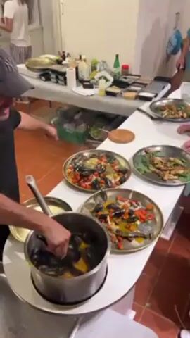Here's a description of the video:

The video takes place in what appears to be a kitchen or catering space.  A man wearing a gray baseball cap and dark clothing is transferring mussels from a large pot into serving plates. The plates already contain fish, tomatoes, and a yellow sauce. The man is using a large spoon to scoop the mussels. There are three plates on the counter in front of him. In the background, there's a long table covered in a white cloth, with various containers, bottles, and other cooking supplies.  Two other people are visible, one in a black shirt and apron standing behind the man with the mussels, and another partially visible in a blue shirt near the right side of the frame. The floor is tiled in a reddish-brown color.