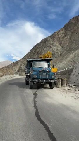 A blue Tata dump truck is parked on the side of a paved road in a mountainous, arid region. The truck is facing the camera, and its dump bed is raised. There is a small puddle of water on the road in front of the truck. In the background, there is a steep, rocky mountain slope. Further in the distance, a white bus is driving away from the camera on the same road. The sky is blue with some clouds. The video pans to show the interior of the dump truck's bed, where people are sitting and there are some orange tarps and bags. The video then pans again to show the bus driving away and then to a wide shot of a valley with a road running through it and mountains in the distance.
