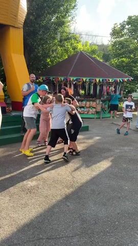 A group of people are gathered outdoors in what appears to be a park or amusement area. In the foreground, the paved ground is dappled with sunlight and shadow. A large, yellow, abstract structure is visible on the left. In the center, a woman in a black and white patterned dress is interacting with a young boy in a light-colored shirt and pink pants, and another young boy in a gray shirt and black shorts. A man in a green shirt and a blue cap is also engaged in this interaction, holding a large bag with a black and white pattern. Other people, including a man in a blue shirt and a woman in a white top, are observing or participating in the background. Behind the central group is a small, red-roofed kiosk with colorful banners displayed. The overall atmosphere seems lively and casual, with several children present.