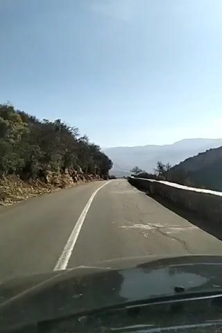 The video shows a view from the driver's seat of a car moving along a two-lane road. The road has a single white line marking the center. On the left side of the road is a rocky hillside covered with trees. On the right side is a low stone wall. In the distance, mountains are visible under a clear, light blue sky. The hood of the car is visible at the bottom of the frame. The road appears to be winding slightly.
