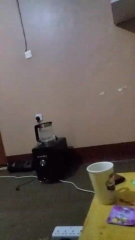 Here is a concise description of the video:

The video shows a panning view of a bedroom. The room contains a bed with a patterned blanket, a shelving unit with various items, a clothes rack with hanging clothes, and a kitchen appliance on the floor. The walls are painted a light color, and there are electrical outlets and a light switch visible. The overall lighting is somewhat dim.