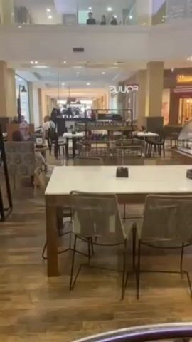 The video shows the inside of a cafe, with tables and chairs in the foreground. The tables are set for customers, with a white marble-like top, wood legs, and wicker chairs. Beyond the cafe seating area, there is a view of a shopping mall hallway with other stores visible. People are seen walking through the hallway in the background. On an upper level of the mall, more people are standing and looking down, appearing to watch the activity below. The flooring in the cafe is patterned wood, and the lighting is bright. The overall scene is calm and unhurried.