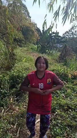 A young girl wearing a red shirt and patterned pants stands outdoors in a natural environment with trees and foliage. She raises her arms in the air and appears to be throwing something, possibly water or powder. She then lowers her arms and crosses them in front of her chest, smiling at the camera. The scene is bright and appears to be taken during the daytime.