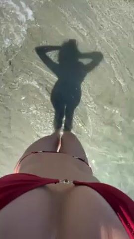 The video shows a view from above of a woman in a red bikini standing in shallow water. Her shadow, dressed in what appears to be a dark wetsuit, can be seen reflected in the water. Gentle waves lap at the shore.
