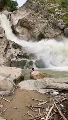 A man stands in a body of water with a large waterfall cascading in the background. The man is partially submerged, with his head and shoulders visible above the water. He is facing away from the camera, towards the waterfall. The waterfall is powerful, with a large volume of water crashing down into the pool. The surrounding environment consists of rocky cliffs and vegetation. The water in the pool appears murky and possibly carries debris.
