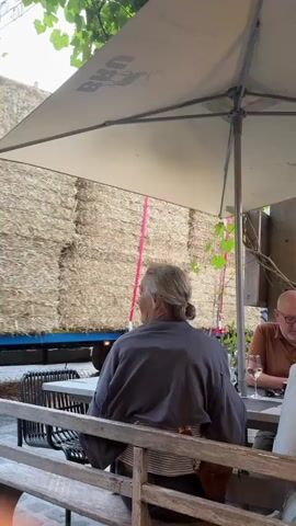 The video shows an outdoor seating area with tables and chairs, some of which are occupied by people. A large truck loaded with hay bales is being driven past the seating area. The hay bales are stacked high on the truck and secured with straps. The truck appears to be moving slowly. The people in the seating area are looking at the truck. The environment appears to be a cobblestone street with some greenery overhead.