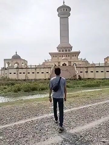 The video captures an outdoor scene featuring a man walking towards a grand architectural complex under a bright, possibly overcast, daytime sky. The man is seen from behind, wearing a light-colored t-shirt, dark trousers, and a dark sling bag across his shoulder. He walks along a gravel path towards the large building in the distance.

The dominant feature of the scene is the impressive, light-colored building complex, which appears to be a mosque or a significant Islamic monument. It features a very tall, cylindrical central tower, reminiscent of a minaret, topped with a dome. This central tower rises from a multi-tiered base. Flanking the main structure are two smaller, symmetrical buildings, each also topped with a dome and featuring arched windows or entrances. A wide set of steps leads up to the main entrance of the central building. The overall architecture is ornate and appears to be constructed from light-colored stone or concrete. The area immediately surrounding the path and the building appears somewhat undeveloped, with patches of green foliage and open ground. No text is visible in the frame. The setting, located in Kruengmate, Indonesia, aligns with the Islamic architectural style observed.