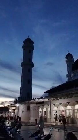 A vertical video shows a mosque at dusk. A tall minaret is on the left, and the mosque building with a dome is on the right. The sky is dark blue with some clouds. The mosque has white walls and arched windows. There are people walking on a paved area in front of the mosque, and several motorcycles are parked in the foreground. The overall lighting suggests it is either sunrise or sunset.