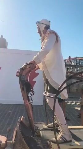 A person in a white costume, goggles, and a hat stands on a treadmill-like device. The person is holding onto a mechanical dragon's head. Another person in a white and red costume joins the first person on the device, also holding onto the dragon's head. They are both moving in sync. The background shows a sunny outdoor setting with a white banner with text and logos. Spectators can be seen in the background. The device appears to be part of a performance or parade.