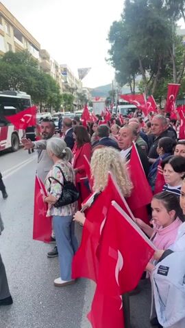 A crowd of people is on a city street waving Turkish flags. There are men in suits, older women, young women, and children in the crowd. Some are standing along the side of the road, while others are moving forward. A bus and several other vehicles are visible in the street. The buildings and trees indicate it is daytime.