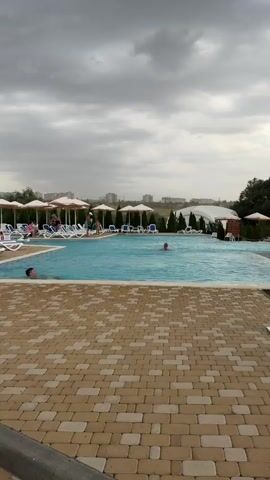 The video opens with a woman standing in front of a swimming pool area. She is wearing a light blue button-down shirt and sunglasses, and she is holding a white hat and a dark blue towel. She smiles at the camera. The scene then shifts to a close-up of a young girl with long, blonde hair, wearing a pink and white striped dress. She is holding someone's hand and laughing as her hair blows in the wind. The video continues to show the girl walking, her hair flowing. In the background, there are people around a pool with umbrellas and lounge chairs. The video ends with the girl and the person holding her hand walking away from the camera towards a path lined with trees and buildings, with other people visible in the distance. The sound in the background consists of the words "Uragan, uragan. Uragan," repeated.