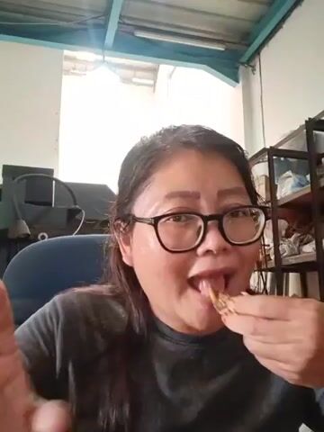 A woman wearing glasses is in the process of eating. She is seated in a chair and the camera is focused on her face. She is holding a piece of food in her right hand and bringing it to her mouth. The background is an indoor setting with a ceiling with metal framing, a window, shelving units with various items on them, and a desk with a computer monitor. The lighting appears to be natural light from the window.