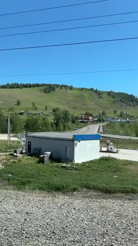 The video shows a rural landscape in what appears to be Russia.  The scene is viewed from a moving vehicle, presumably a train, judging by the power lines visible overhead. In the foreground, there is gravel and green grass.  Several small, rundown buildings can be seen, including a white prefab structure and several brick or concrete buildings.  A road crosses a small bridge. In the background, a grassy hill rises towards the clear blue sky.  Scattered trees dot the landscape and a few houses are visible on the hill.  The overall impression is one of a simple, rural setting.